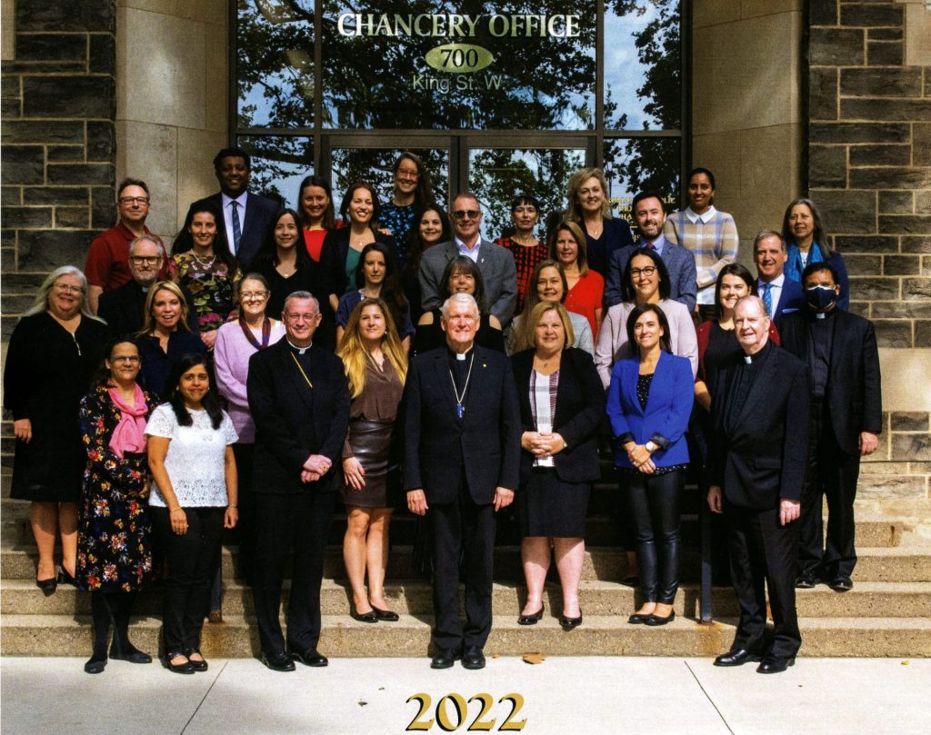 Group photograph of staff from the Diocese of Hamilton Chancery Office in 2022. Staff members stand on the stairs in front of the Chancery building.
